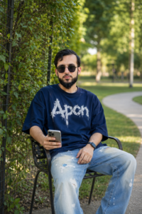 A fit young South Asian man wearing a navy polo sweater and white prayer cap sitting on the hood of a black SUV during golden hour sunset.